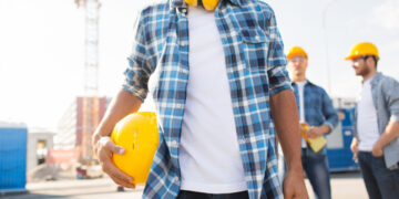 building, construction, protective gear and people concept - close up of builder holding yellow hardhat or helmet outdoors